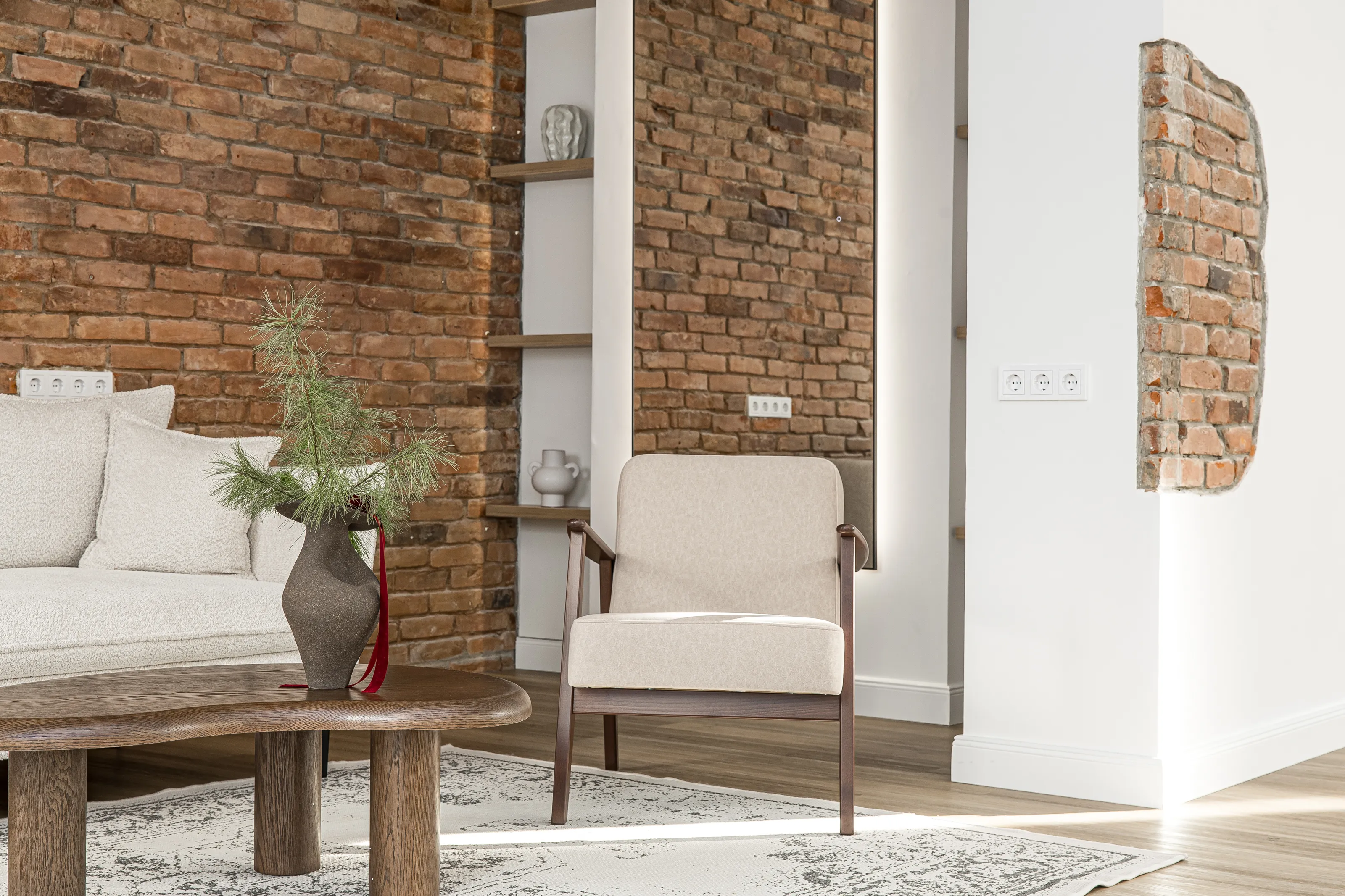 Fotelis prie plytų sienos su dekoratyviniu plytų fragmentu ir lentynomis